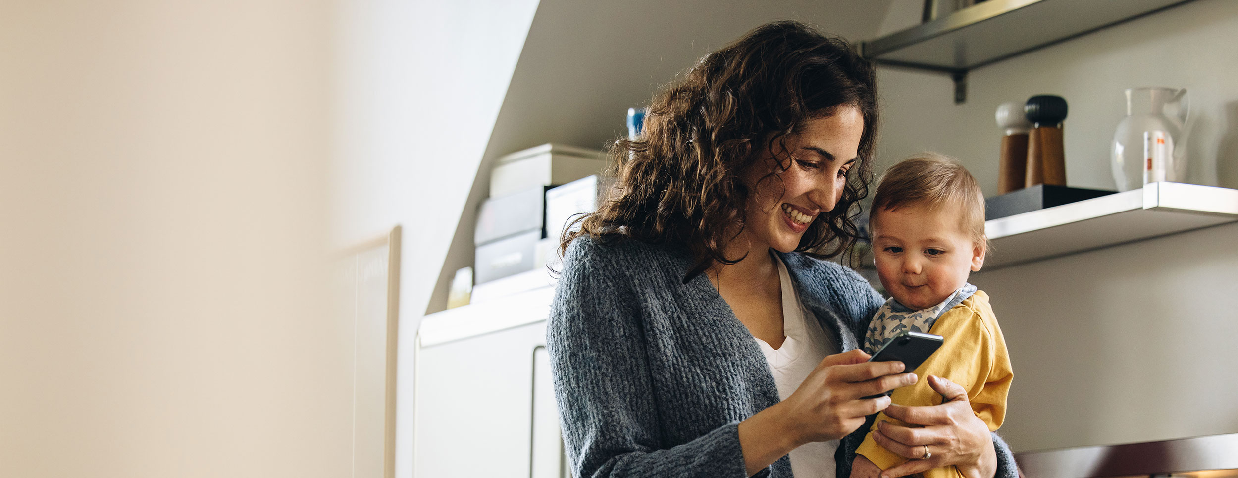 A mother smiling and holding her baby as she looks at her phone